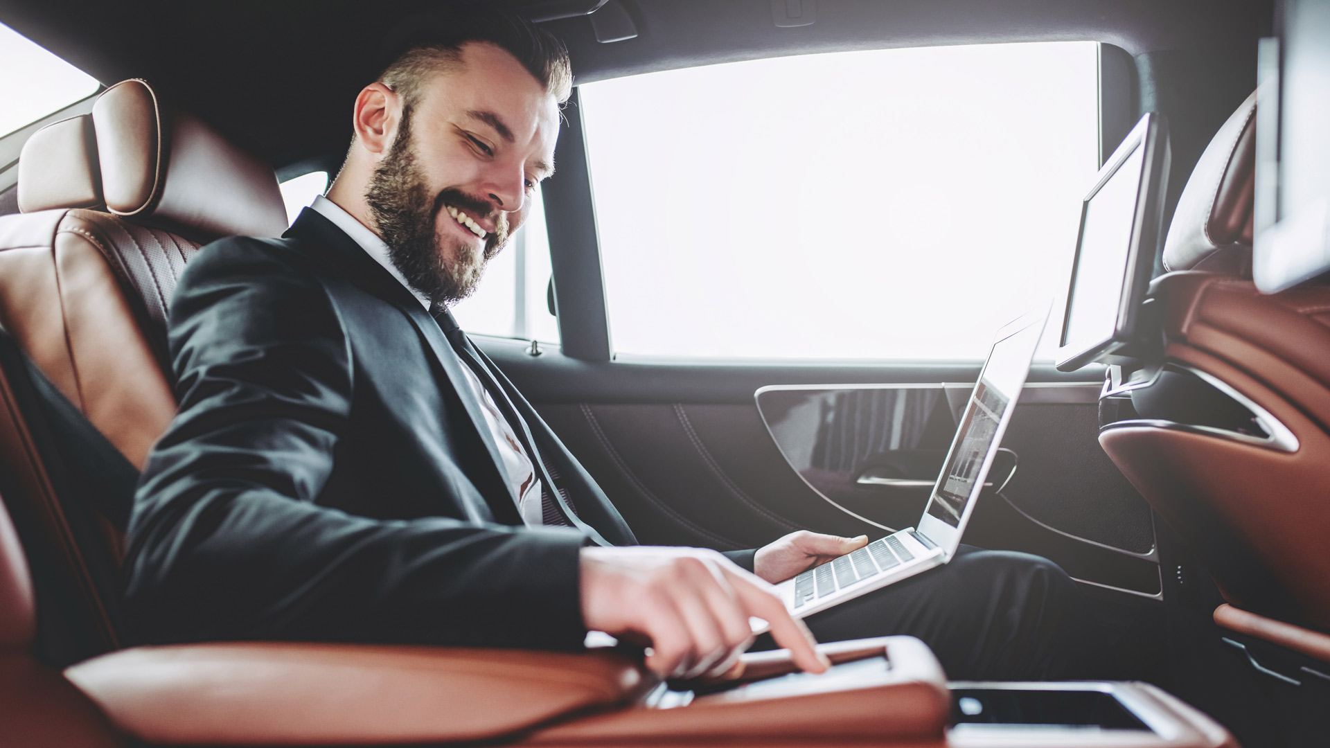 Young handsome businessman is sitting in luxury car. Serious bearded man in suit is working with laptop while being in trip.